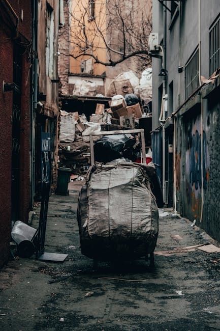 A narrow urban street scene shows several workers in high-visibility vests and protective helmets gathered near a large, open-top black rubbish bin, which is filled with assorted waste materials, incl